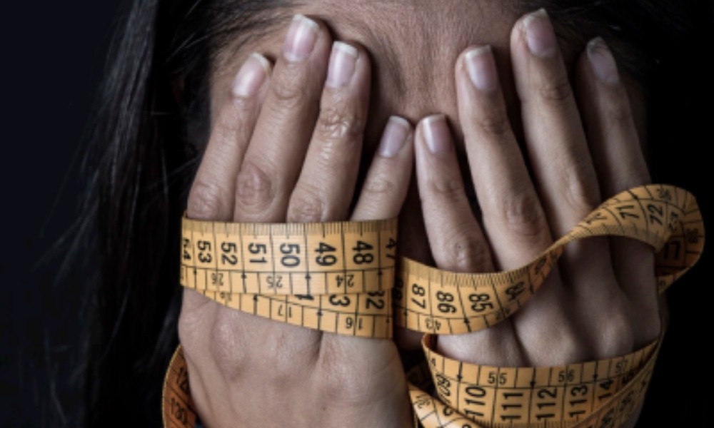 Person covering face with hands wrapped in a measuring tape, symbolizing body image or weight concerns.