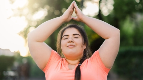 Person in athletic clothing standing outdoors with arms raised overhead in a yoga stretch.